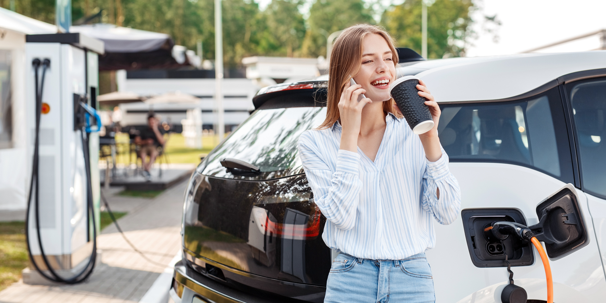 Billede af kvinde, som er i gang med at oplade sin elbil ved en rasteplads, mens hun snakker i telefon og drikker en kop kaffe.